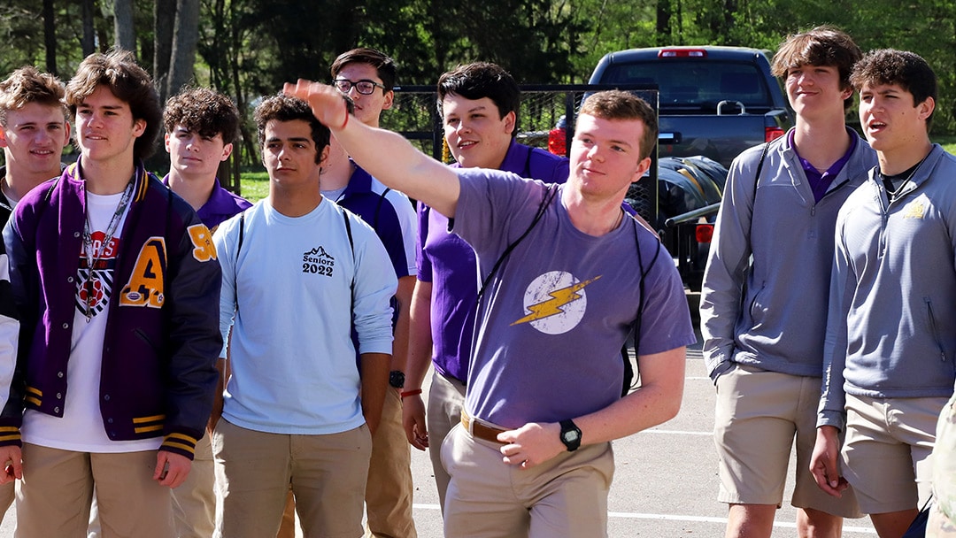 A group of high school boys dressed in purple school colors watch as a classmate throws a baseball towards a target
